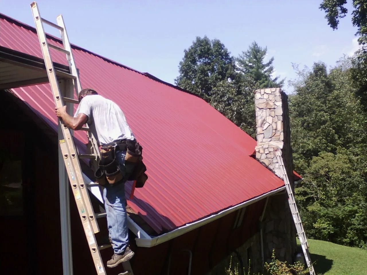 Roofer on a ladder beside a red metal roof and stone chimney — Soffit & Fascia Repair by Roofing Brothers in Melrose